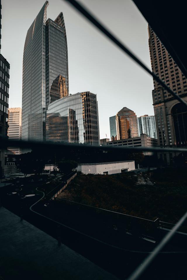 Tall glass office buildings in a modern city center reflecting soft daylight, seen through a barrier, capturing the structured, reflective spaces where blending in with neutral clothing and average behavior supports urban anonymity. Modern downtown office towers viewed through a fence, showing an urban setting where wearing low-profile, gray man gear allows preppers to stay unnoticed and move strategically during crisis scenarios.