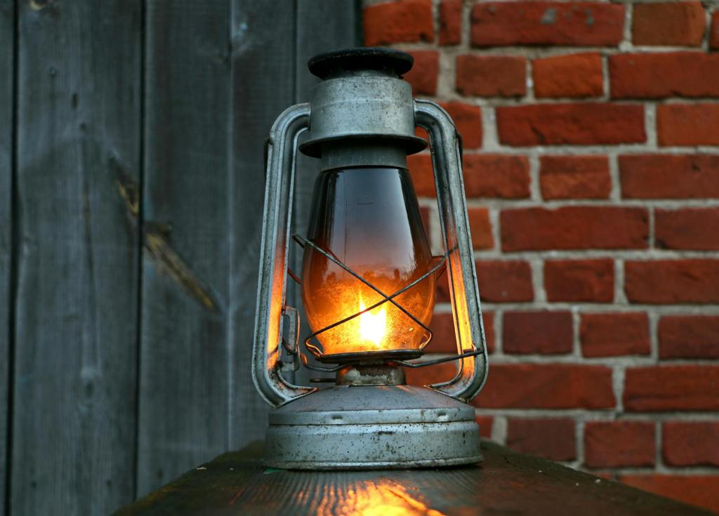 Oil lamp on wooden surface with bricks and wooden fencing behind, representing analog light for self-reliance.